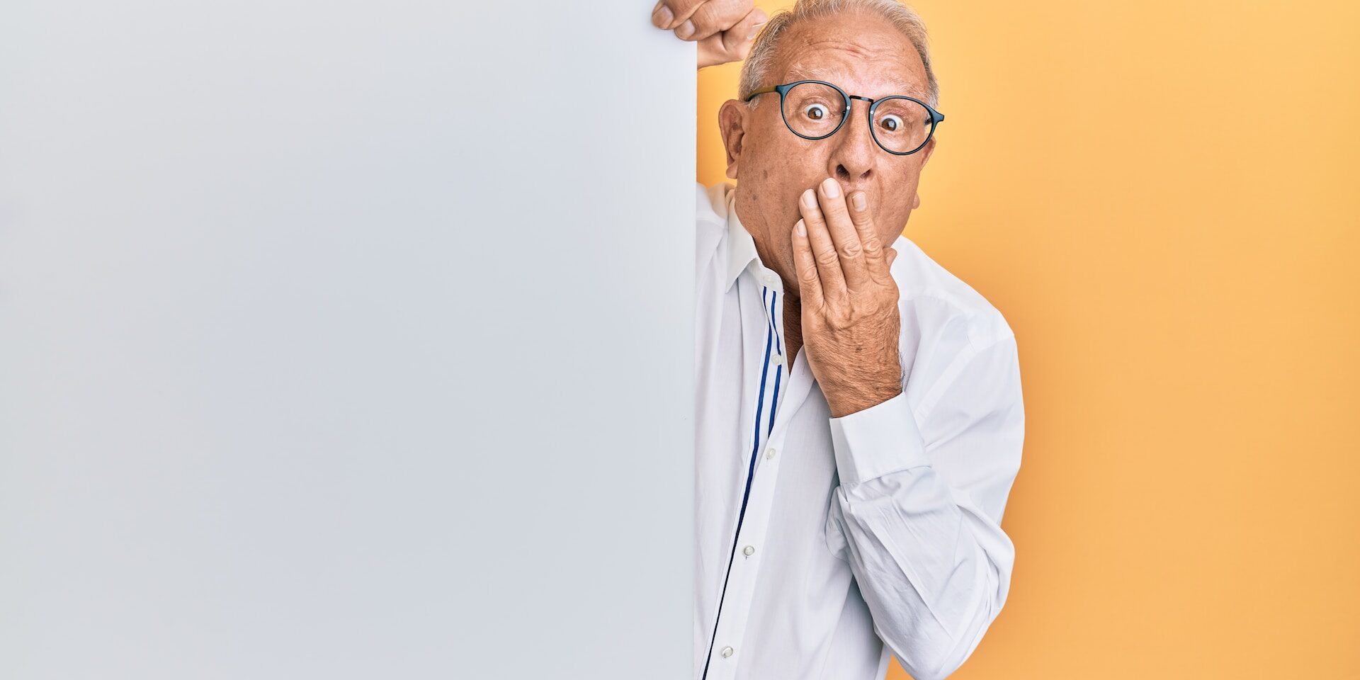 older man in white dress shirt peeking out from behind white door, covering mouth in front of orange wall