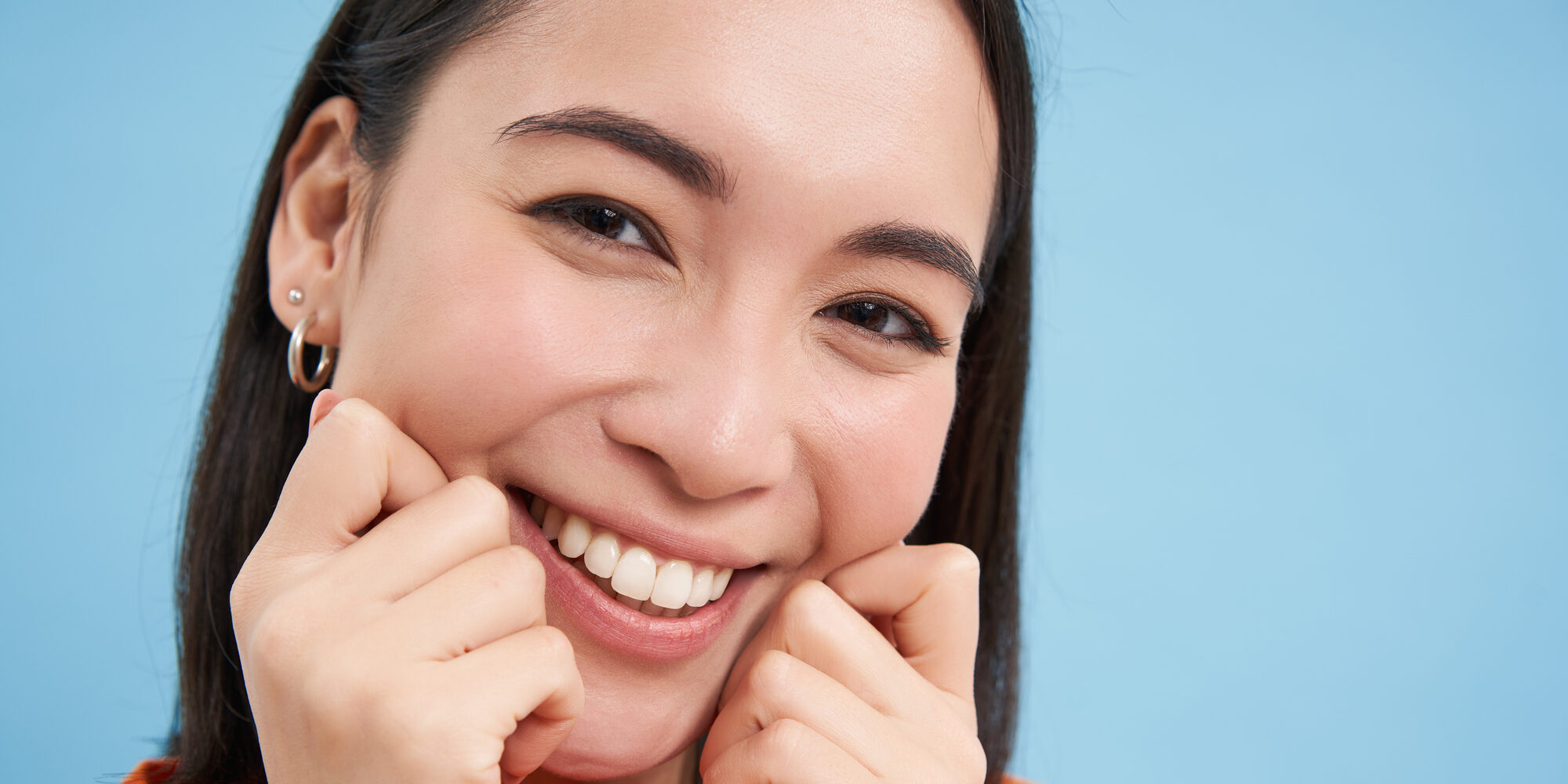 woman with bright white teeth pinching her cheeks and smiling on blue background