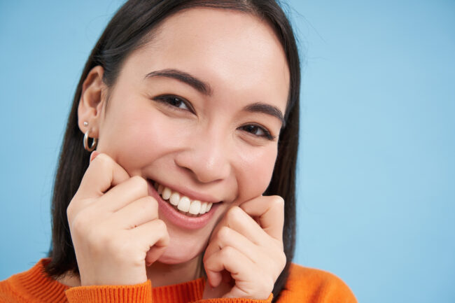 woman with bright white teeth pinching her cheeks and smiling on blue background
