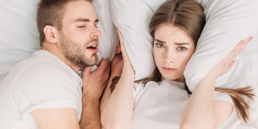 man snoring, laying next to wife, wife covering her ears with a pillow