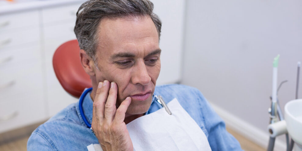 older man sitting in dentist chair, touching face because of tooth pain