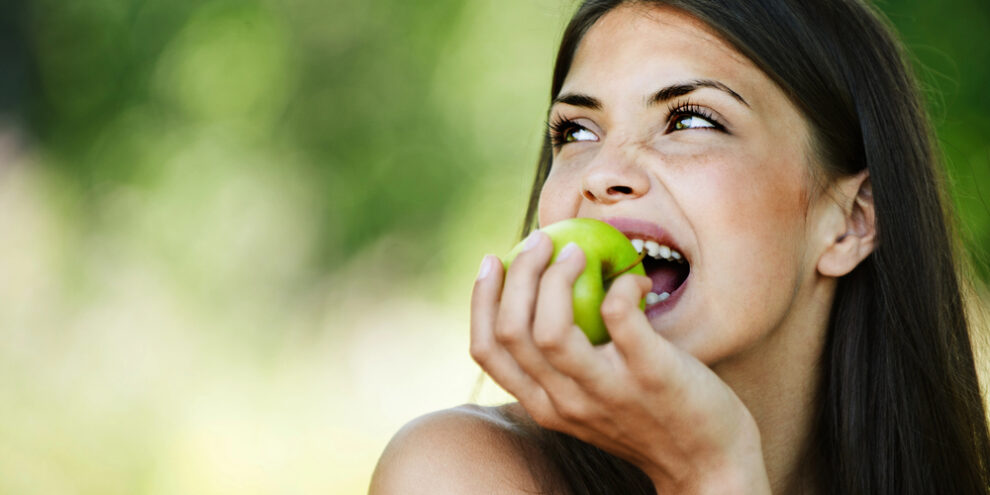 young woman smiling, looking away from camera, and eating an apple