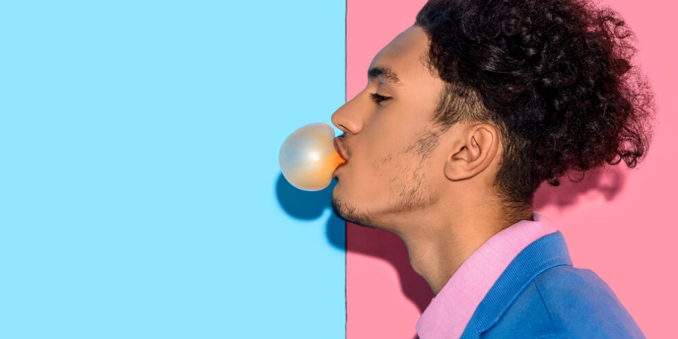 young man blowing a bubble gum bubble against a split background of soft pink and blue