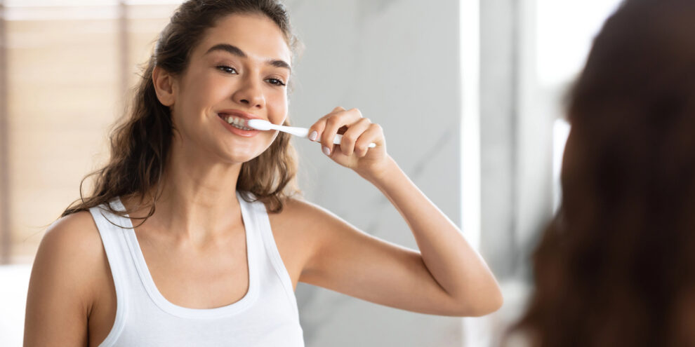 woman with brown curly hair brushing her teeth in bathroom mirror